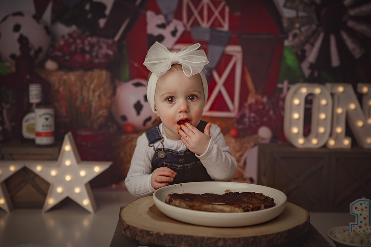 A happy baby sits in a barn themed studio eating a steak in denim overalls and large white bow