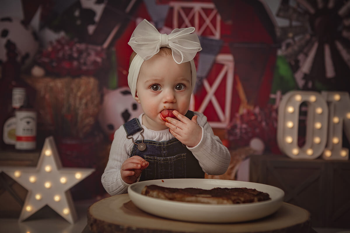 A baby girl in denim overalls and a big white bow licks her fingers while enjoying a steak in a farm themes studio before visiting a corn maze in springfield, il