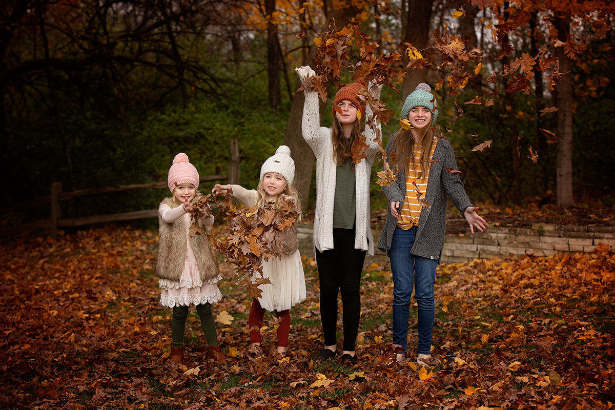 Happy cousins toss leaves in the air above themselves while standing in a lawn in sweaters and beanies