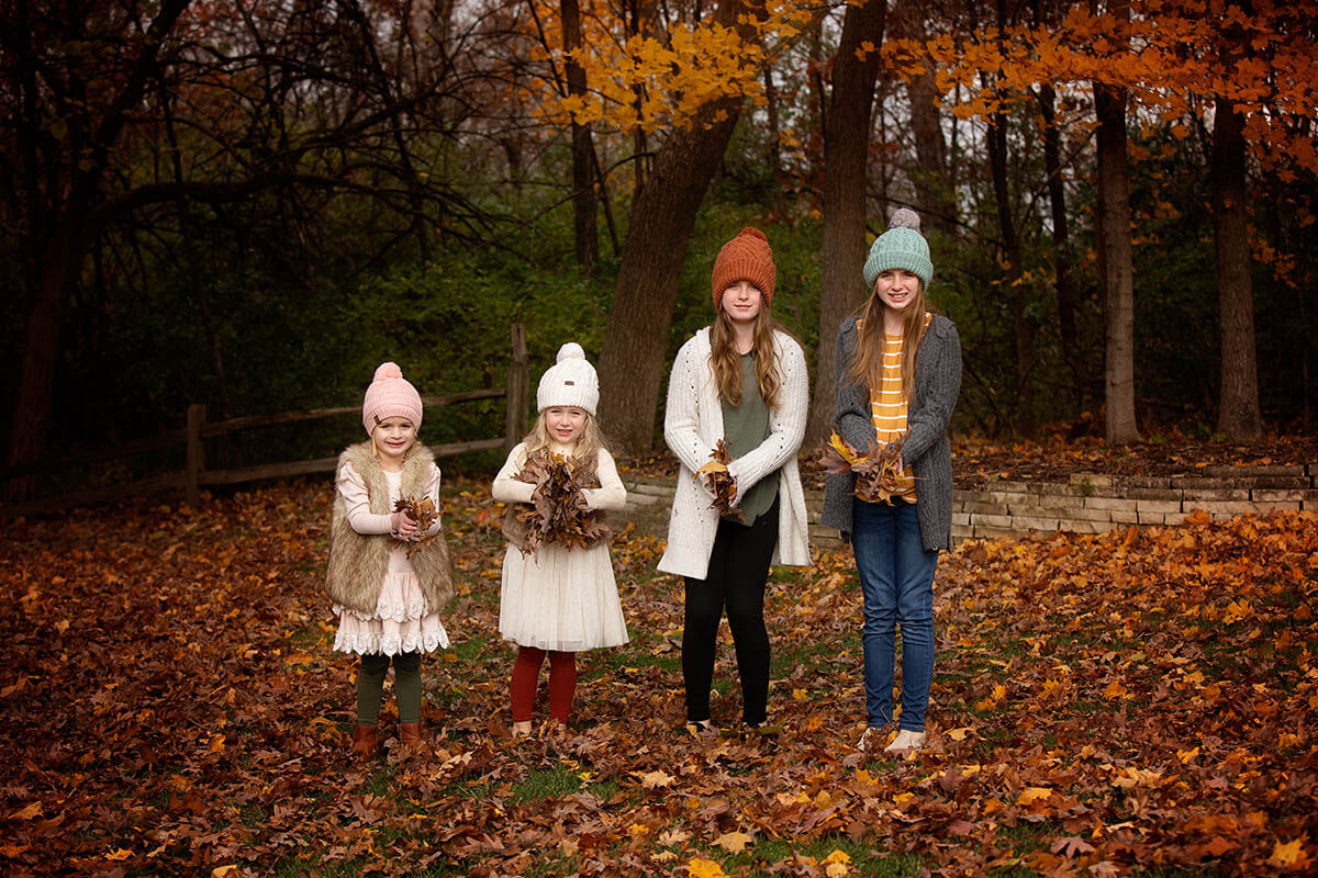 Four young cousins collect leaves in a lawn in fall while wearing sweaters and white dresses