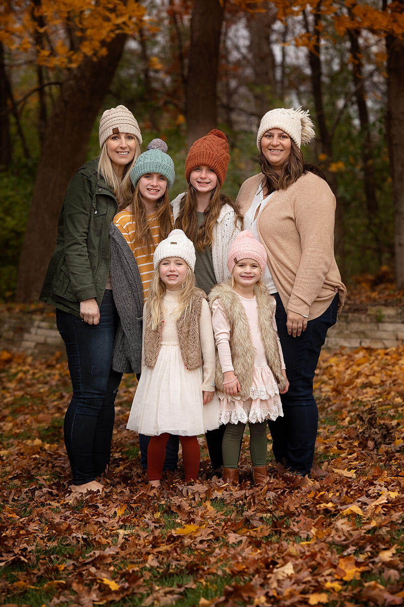 Two moms in sweaters stand with their four daughters in a lawn covered in fall leaves with all in beanies before some fall activities in Springfield, IL