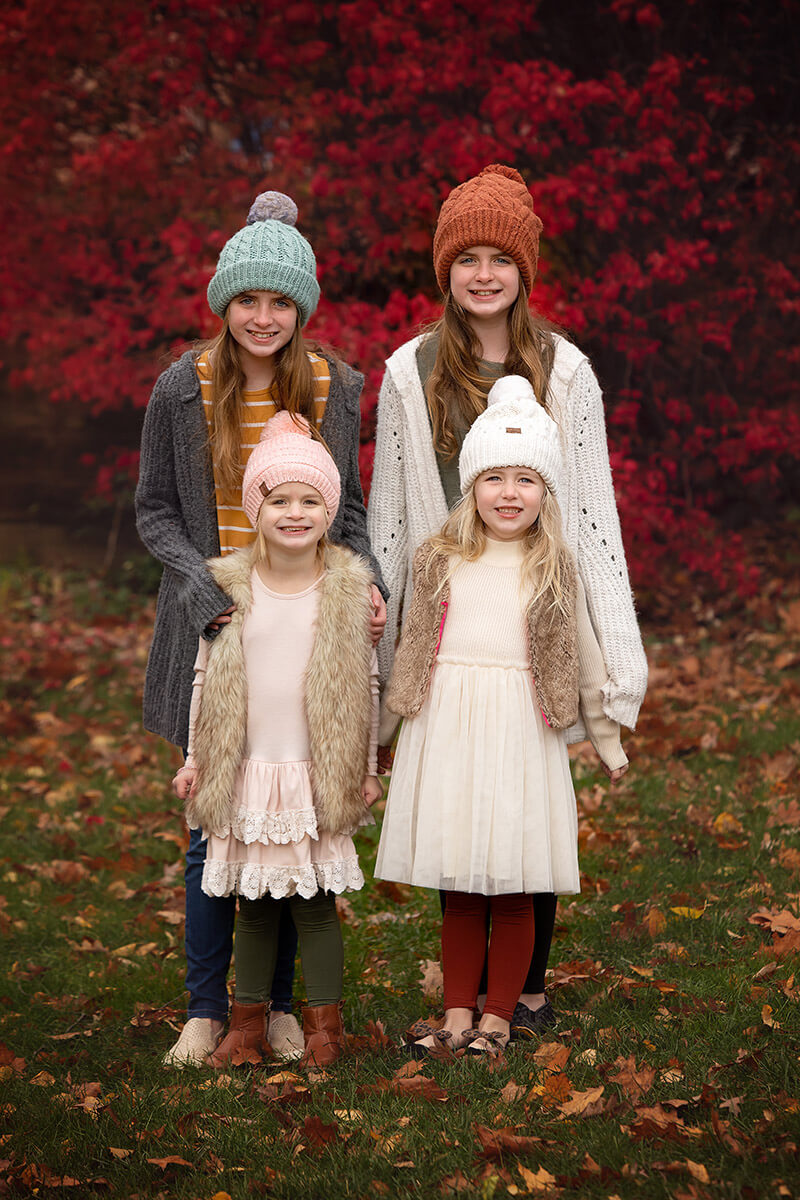 Four young cousins stand in beanies and sweaters in a lawn covered in fallen leaves before some fall activities in Springfield, IL