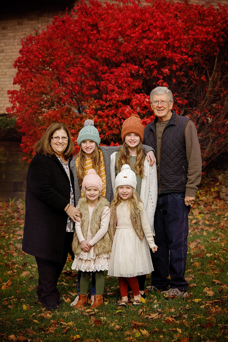 Smiling grandparents stand with their four granddaughters in front of a bright red bush before visiting fall festivals in Springfield, IL