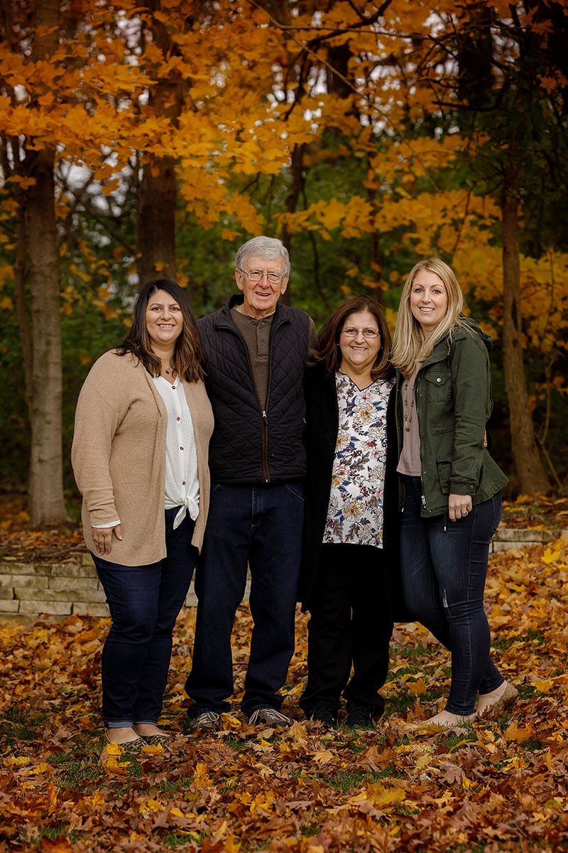 Smiling parents stand with their happy adult daughters in a lawn covered in fall leaves