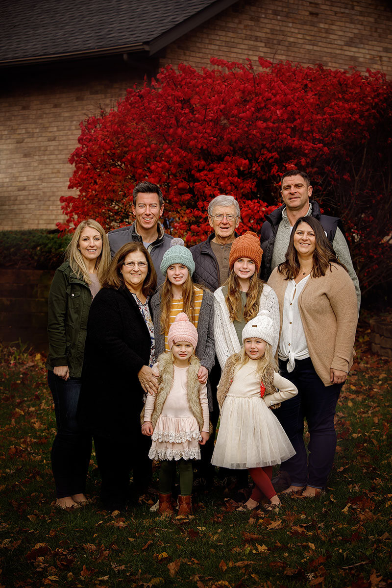 A happy extended family with 3 generations stand in front of a bright red bush in cozy outfits before visiting fall festivals in Springfield, IL