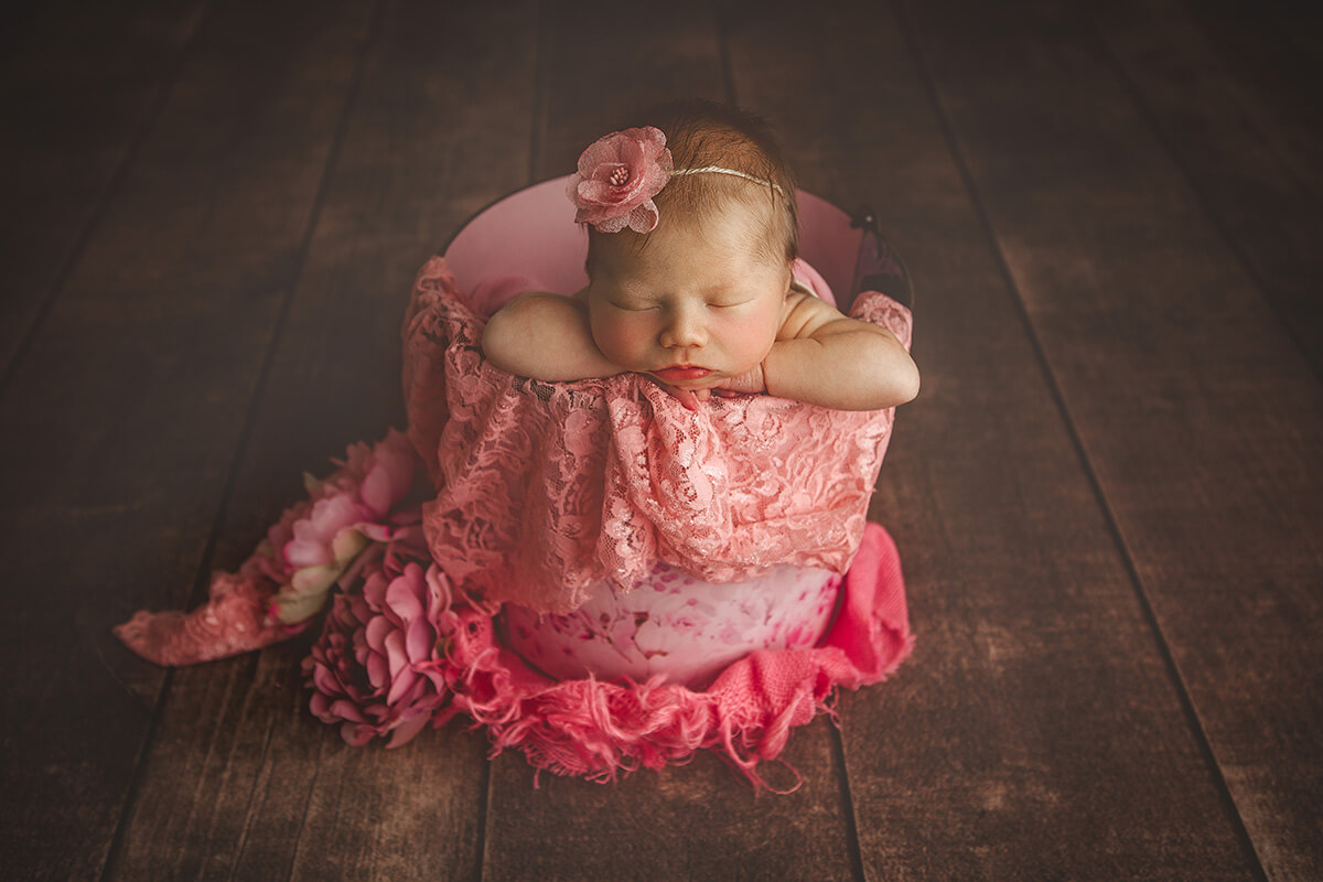 A sleeping newborn baby leans on the rim of a pink bucket in studio