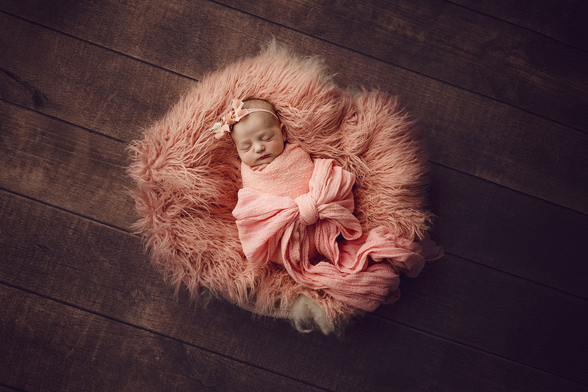 A sleeping newborn baby wrapped tight in a pink swaddle on a fur blanket on wood floor after meeting an obgyn in Springfield, IL
