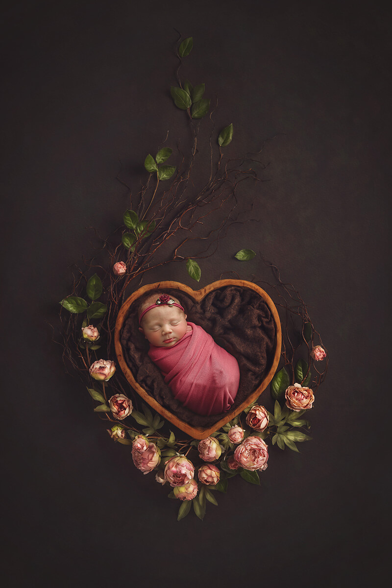 A newborn baby girl sleeps in a wooden bowl surrounded by pink flowers in a pink onesie thanks to an obgyn in Springfield, IL