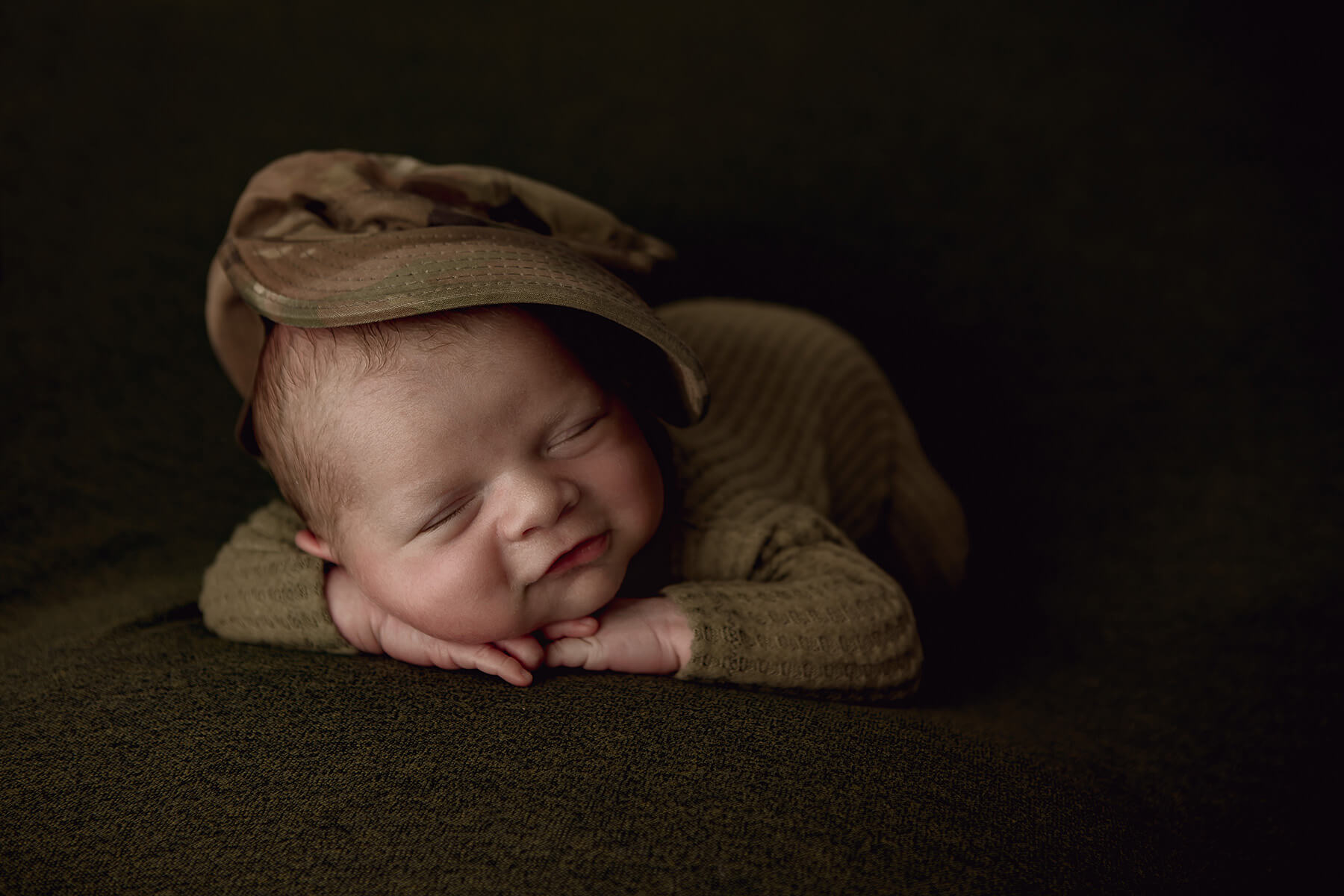 A newborn baby sleeps in a camo onesie with matching hat in froggy pose in a studio