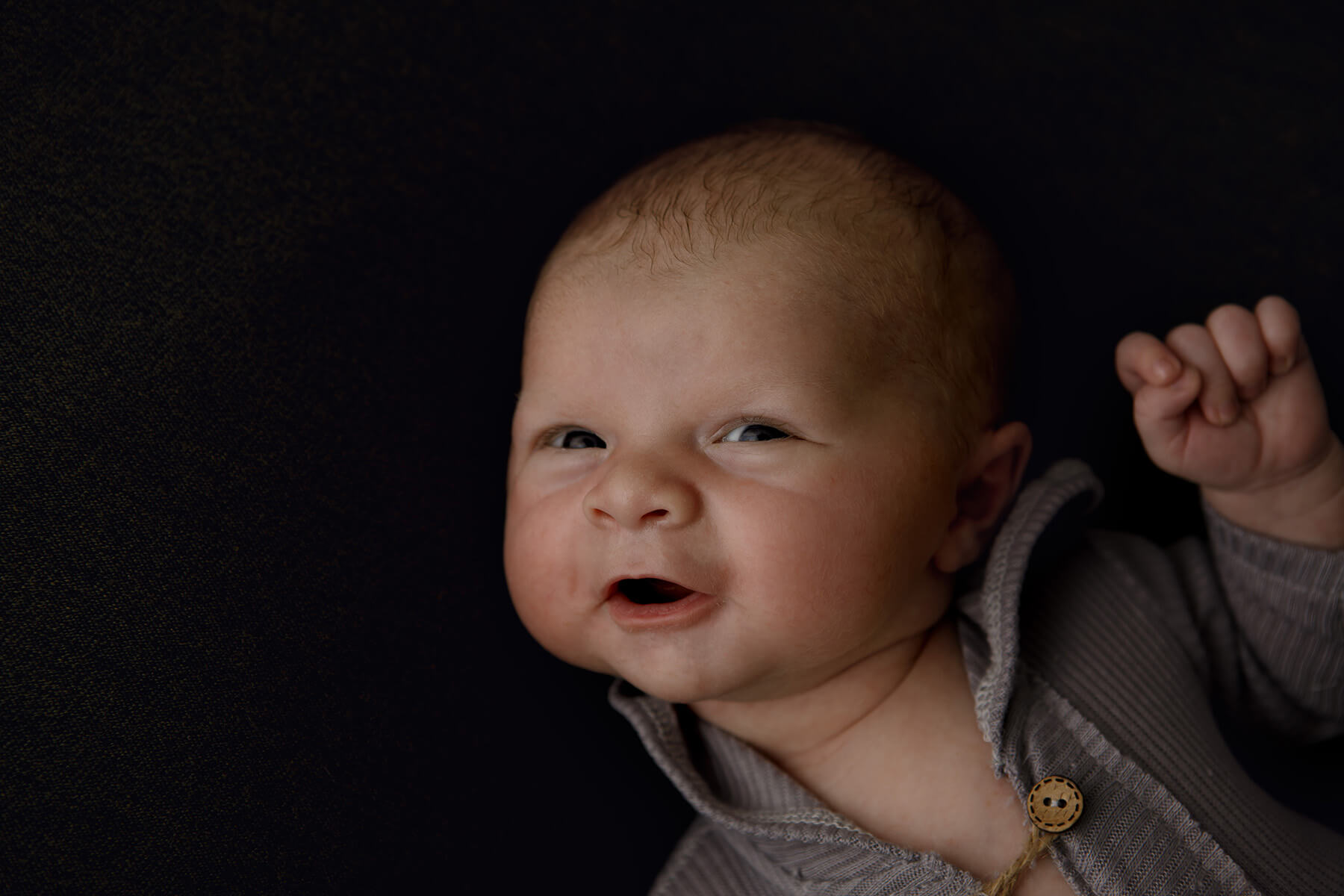 An infant in a grey onesie with eyes and mouth open in a studio