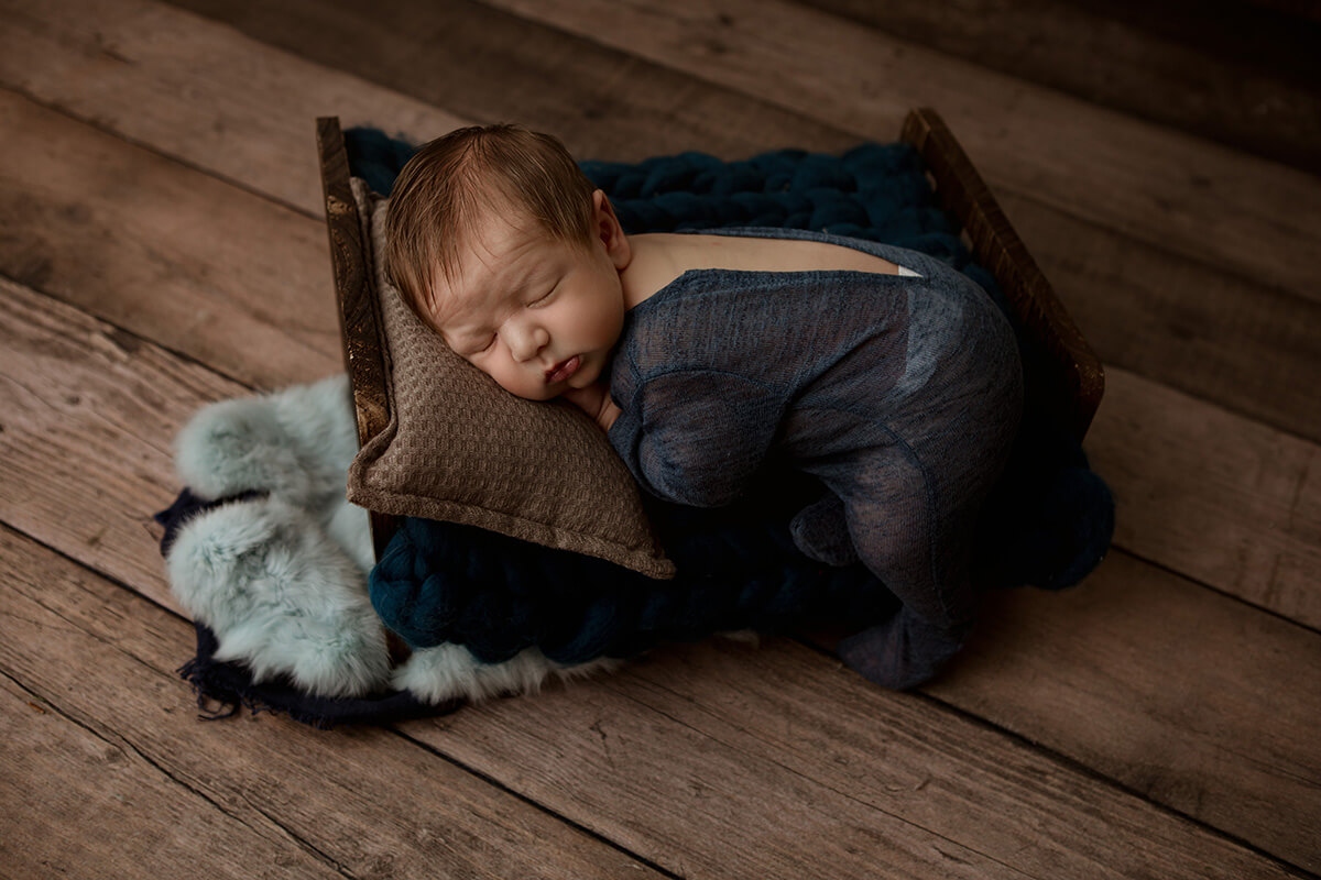 A newborn baby in a blue onesie sleeps on a tiny wooden bed in a studio after mom did prenatal yoga in springfield, il