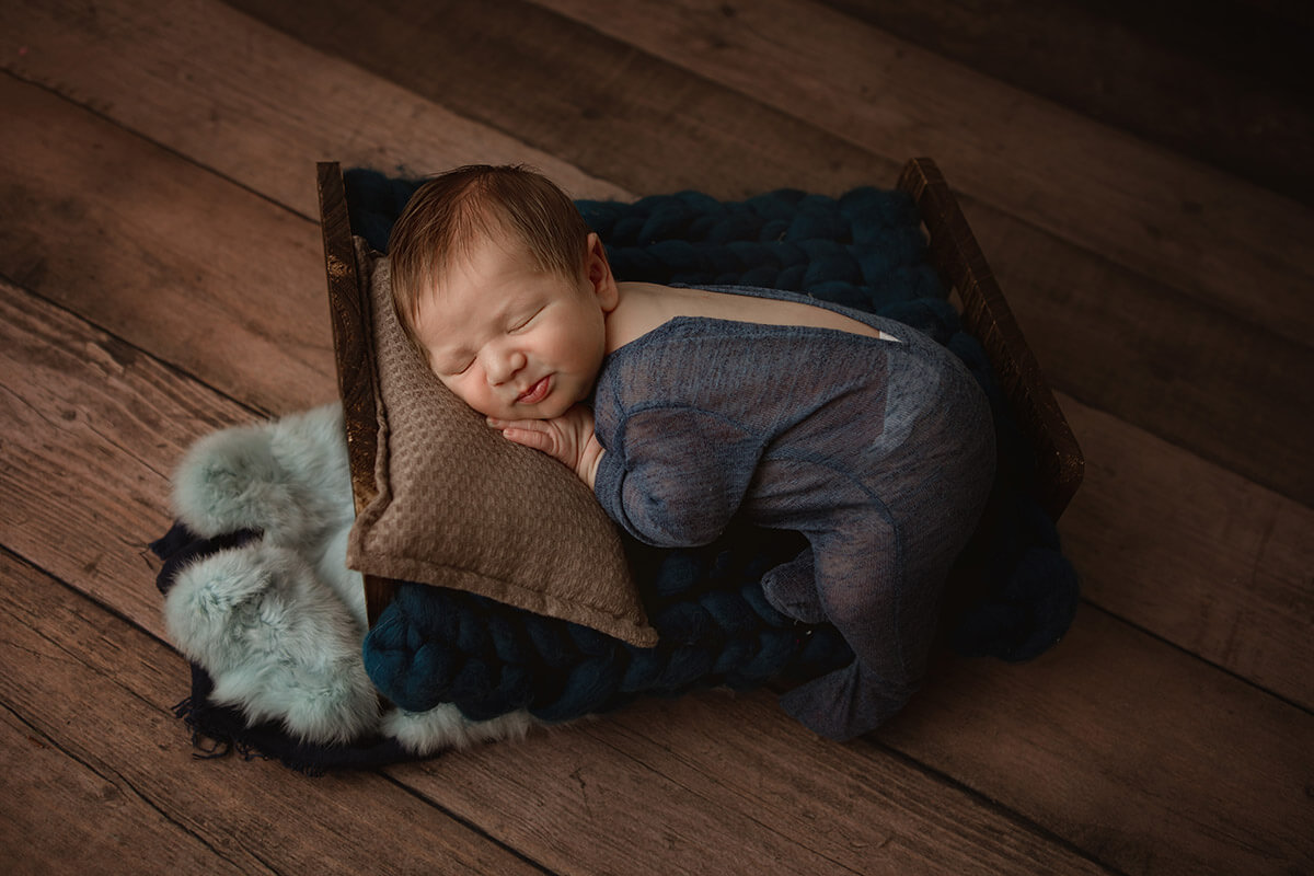 A sleeping newborn in a blue sheer onesie sleeps on a wooden bed on wood floors in a studio before finding tongue tie specialists in Springfield, IL