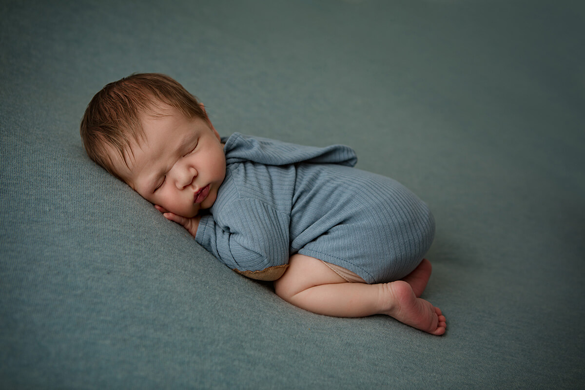 A sleeping newborn baby in froggy pose in a blue onesie with a hood in a studio before finding tongue tie specialists in Springfield, IL