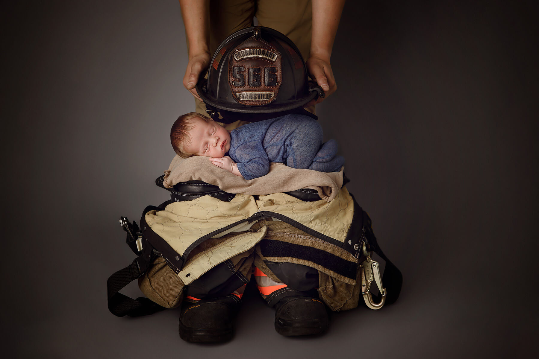 A newborn baby sleeps on a stack of fireman gear under the probationary helmet held by dad before finding tongue tie specialists in Springfield, IL