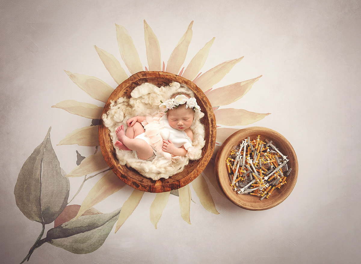 A newborn baby sleeps in a wooden bowl in a white flower dress next to a bowl of IVF syringes