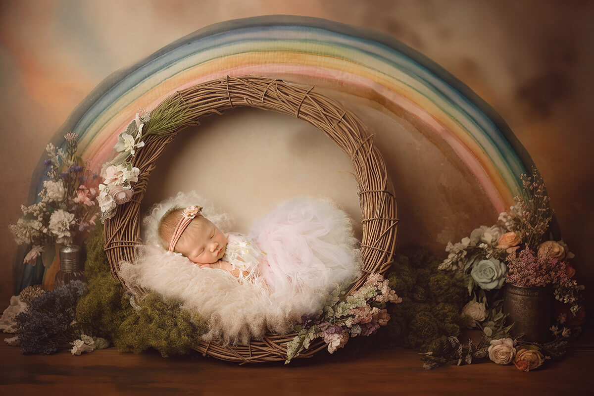 A sleeping newborn girl in a pink and white gown on a wicker wreath under a rainbow thanks to birth center of bloomington normal
