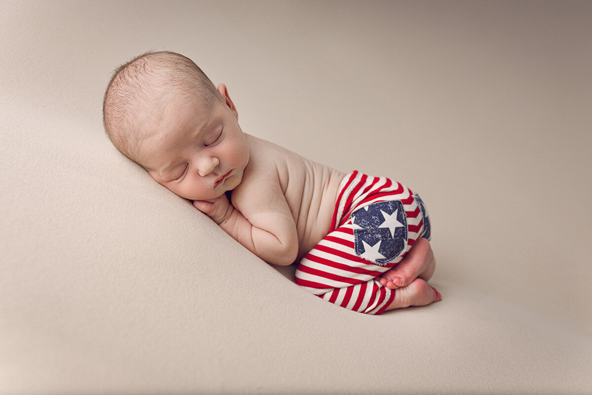 A newborn baby in american flag pants sleeps in froggy pose in a studio thanks to birth center of bloomington normal