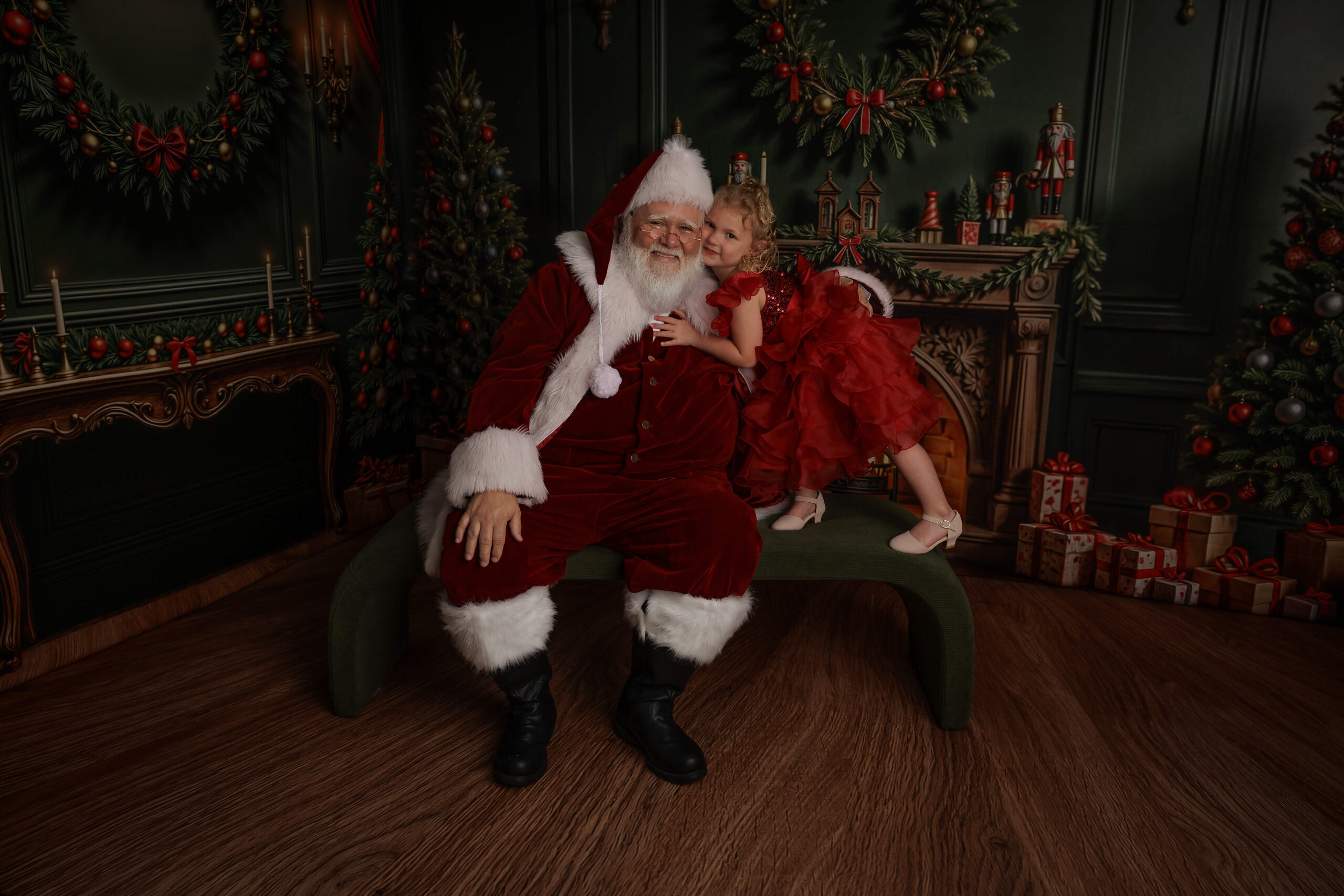 A happy Santa sits on a green bench in a living room hugging a toddler girl in a red dress