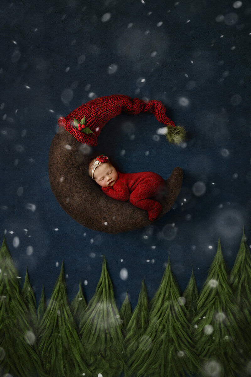 A sleeping newborn on a moon in a red onesie while it snows