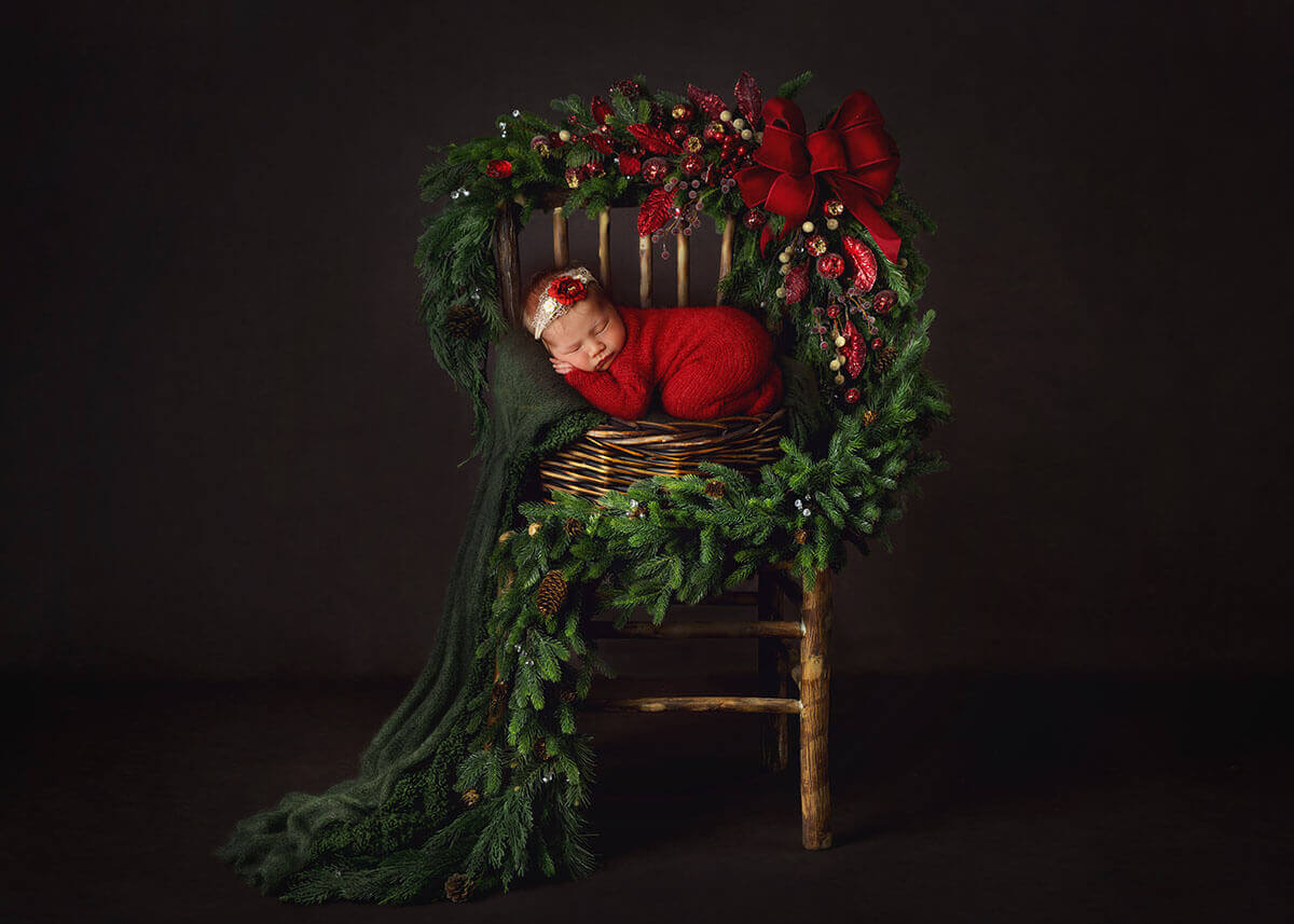 A sleeping baby girl on a tall wicker chair draped in garland in a red knit onesie after finding christmas lights in springfield, IL