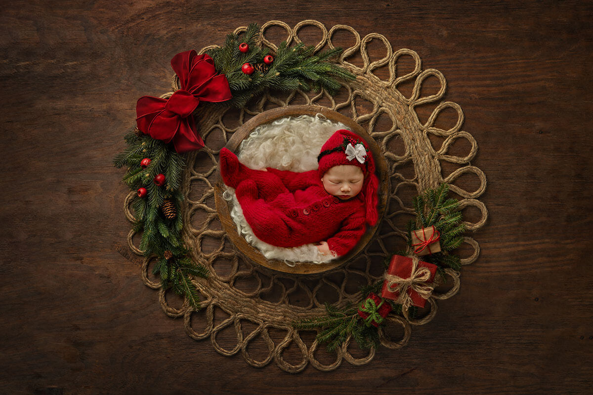a newborn baby sleeps in a wooden bowl surrounded by greenery wearing a red onesie and matching santa night cap after visiting christmas lights in springfield, IL