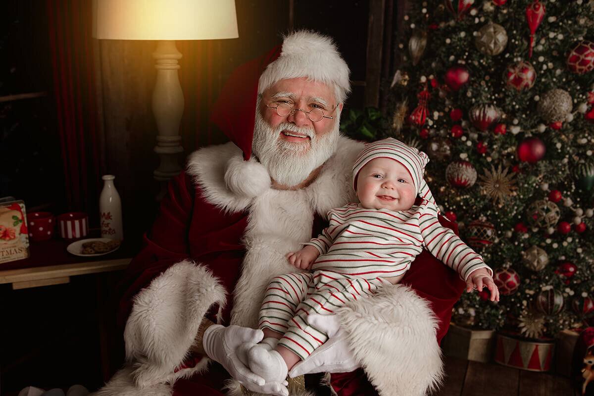 A smiling santa sits in front of a tree with a happy baby in stripe pajamas in his lap