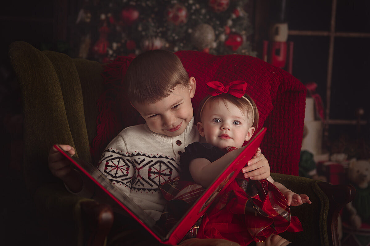 A young boy in a white sweater reads a book in a chair to his baby sister sitting in his lap in a red dress before some christmas parades in springfield il