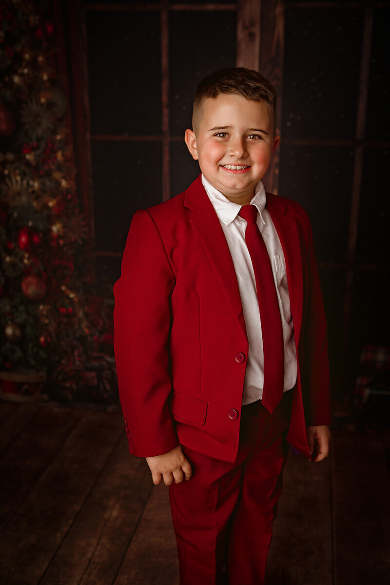 A smiling young boy in a red suit and tie stands in a studio with hands by his side before attending christmas parades in springfield il