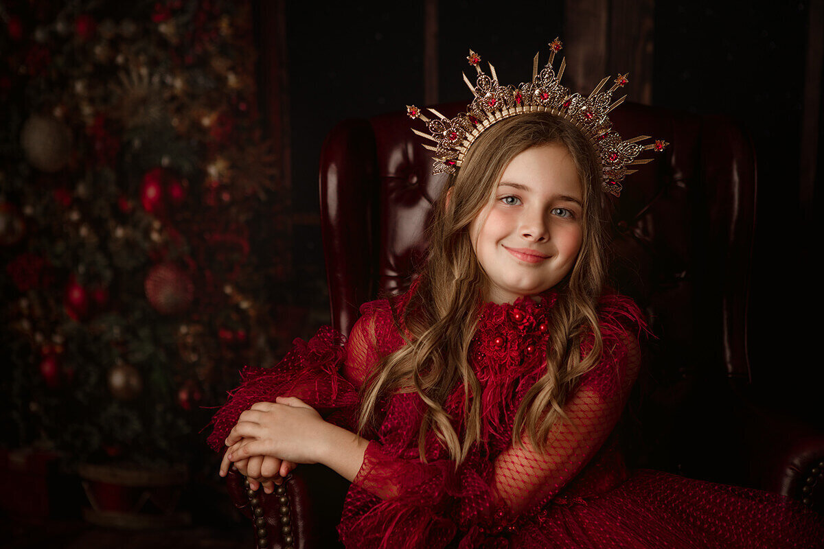 A smiling young girl in a red gown leans on the arm of a vintage leather chair in a studio in a large tiara before watching christmas parades in springfield il