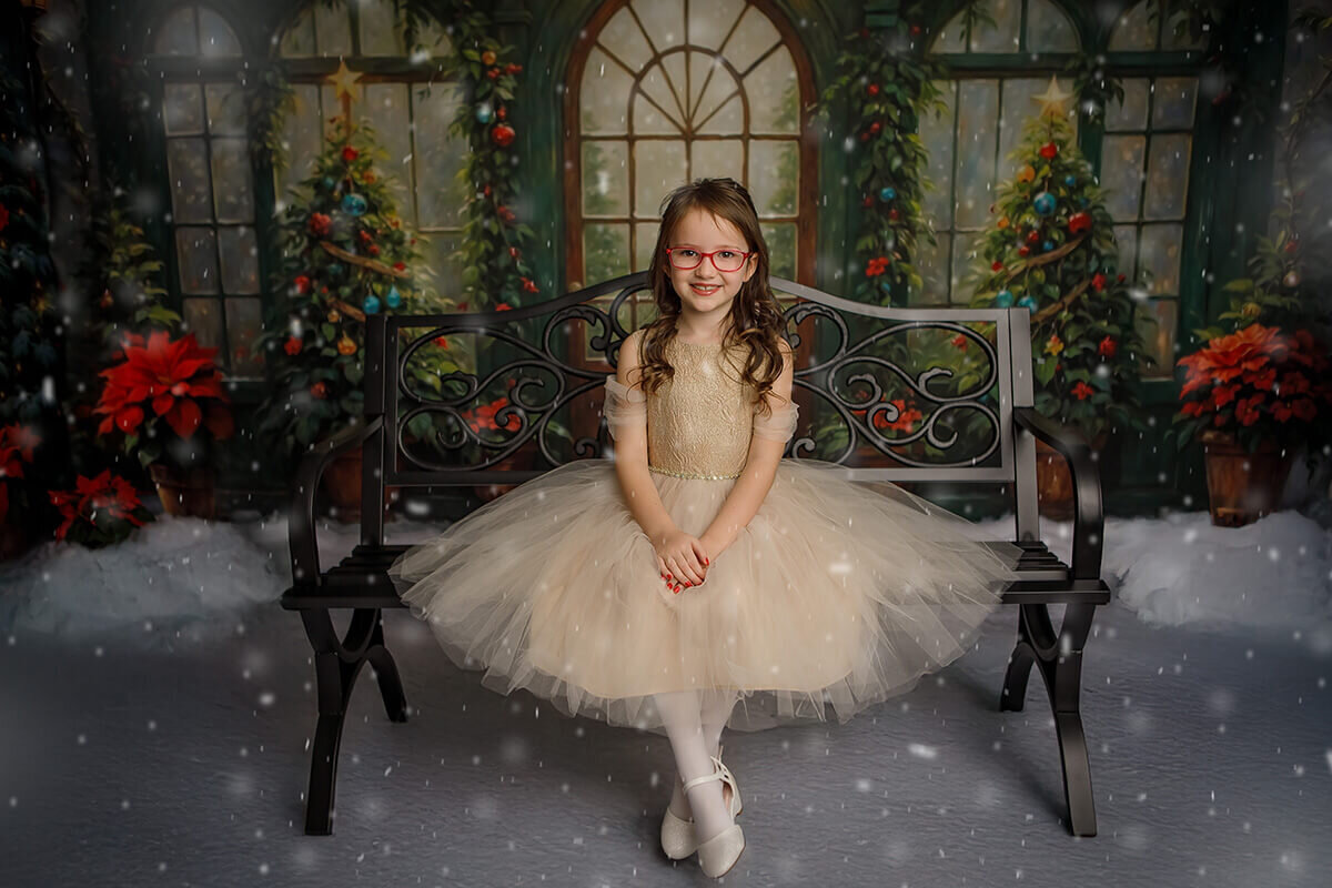 A happy young girl in a gold gown sits with hands in her lap on a bench in a studio with snow falling