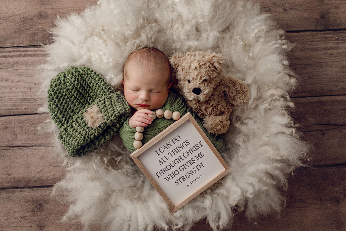 A sleeping newborn holding a wooden sign in a green swaddle with a teddy bear on a furry blanket