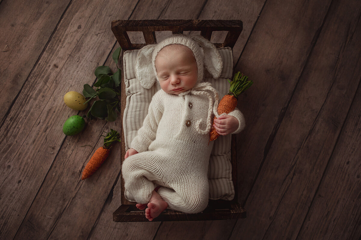 A sleeping newborn cuddling a knit carrot in a white knit bunny onesie in a tiny wood bed