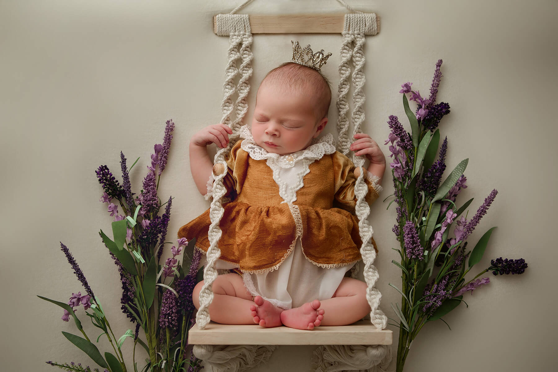 A newborn baby girl sleeps while sitting on a swing ina. studio surrounded by lavender in a gold dress before visiting daycares in bloomington il