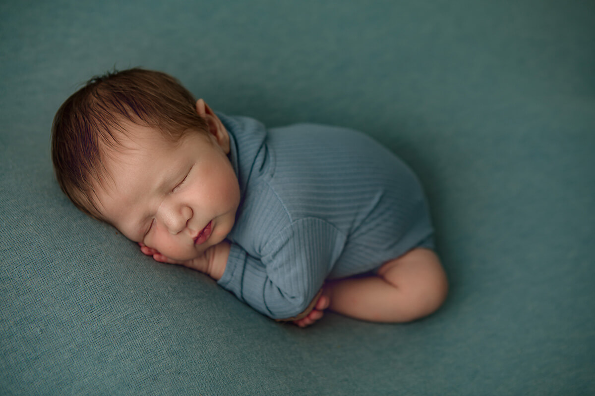 A sleeping newborn in a blue onesie sleeps on his tummy in froggy pose in a studio