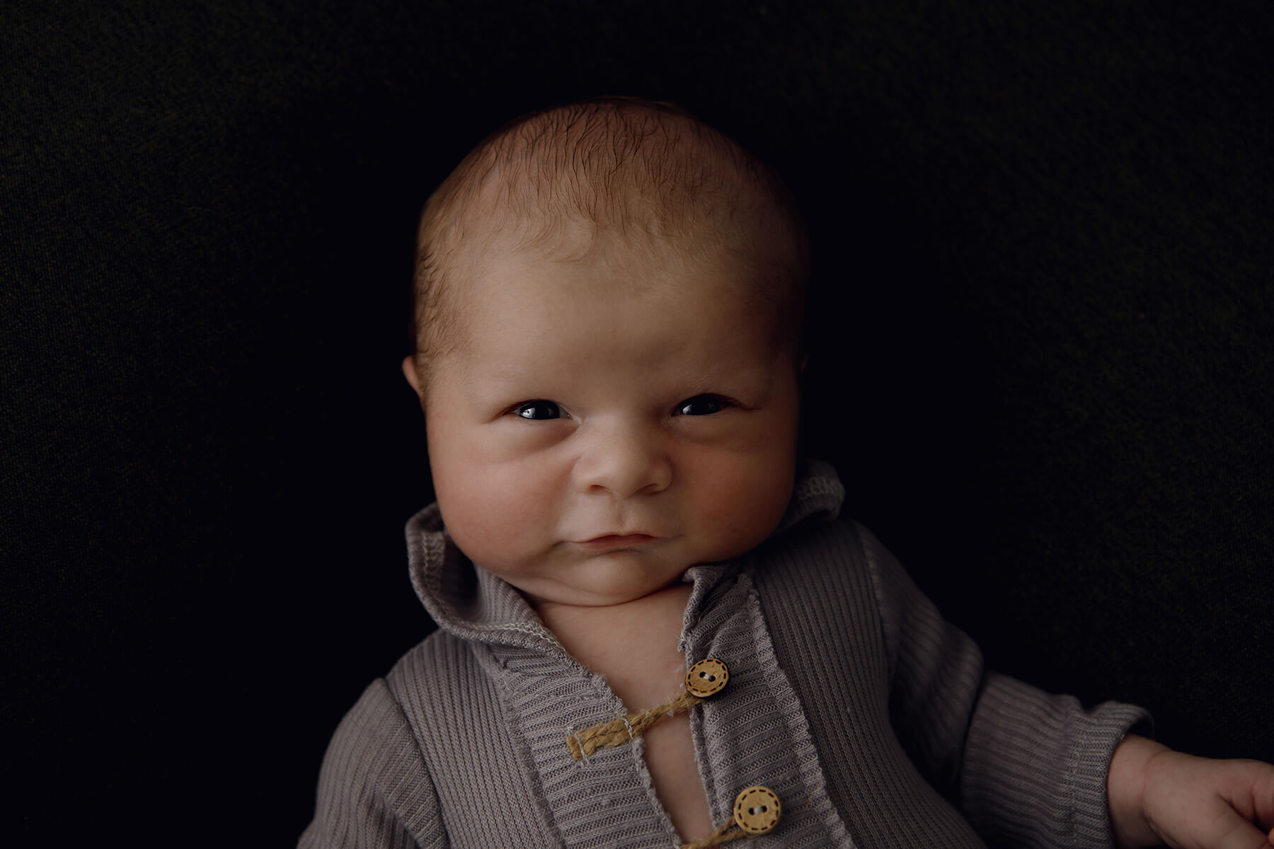 A newborn baby lays on a black bed in a grey onesie with eyes open and a squish face