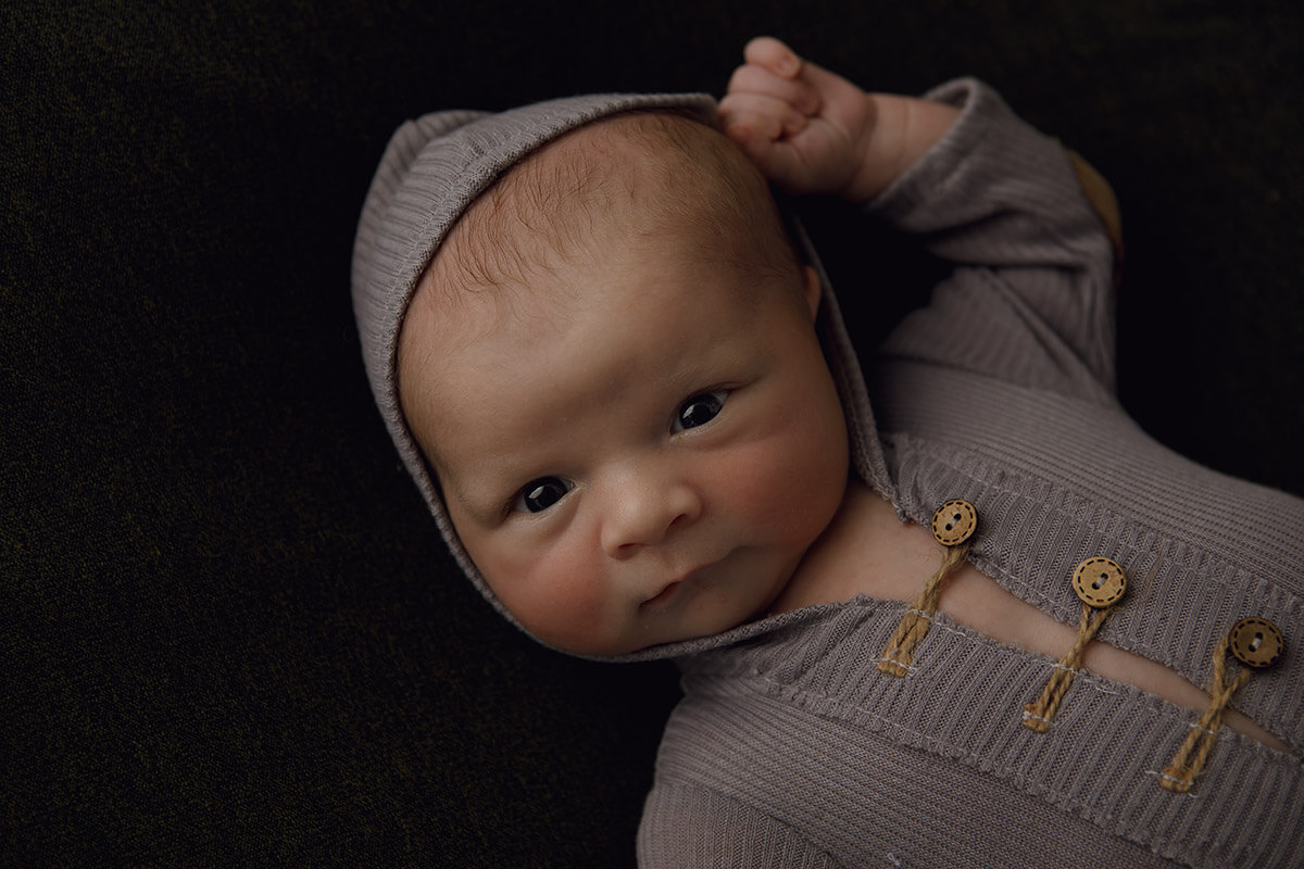 A newborn baby lays in a brown onesie with a hood with eyes open and an arm up thanks to doulas in bloomington il