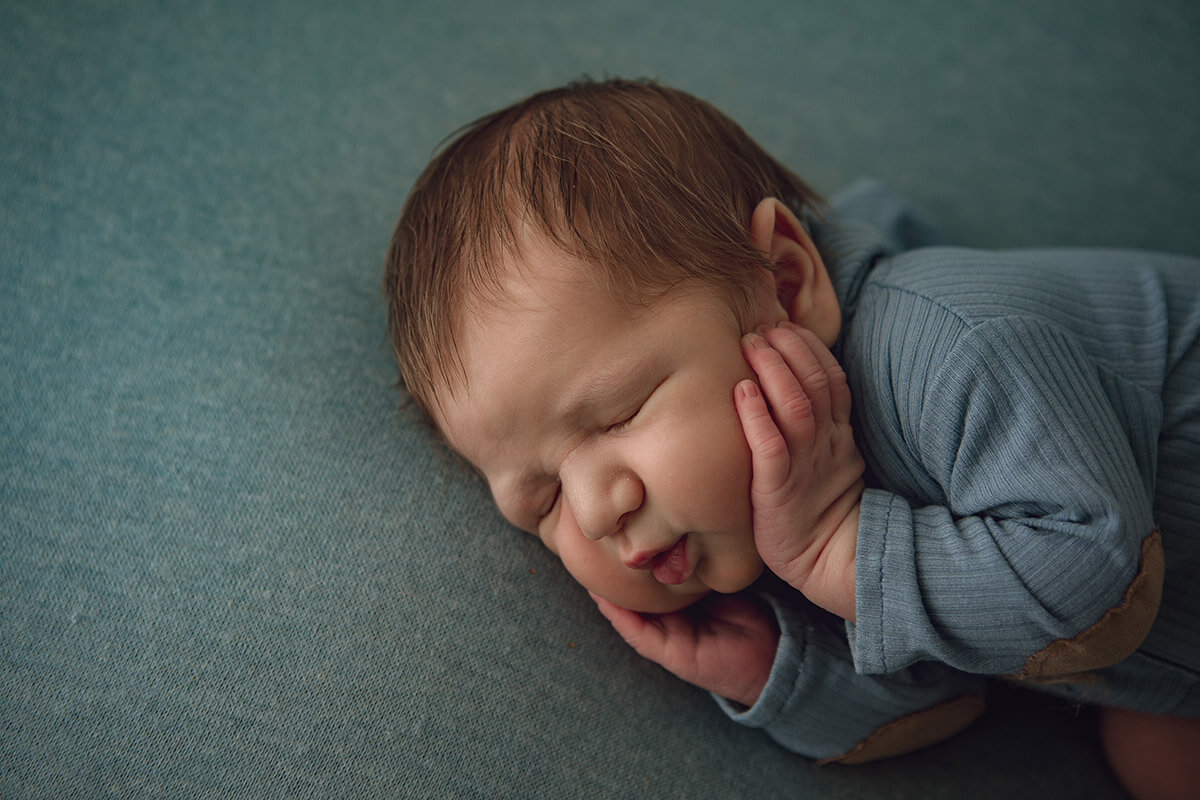 A newborn boy holds his hands on his cheeks while sleeping in a blue onesie on his side after meeting doulas in bloomington il