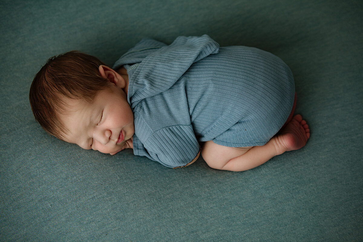 A sleeping newborn baby boy in a blue onesie sleeps in froggy pose after meeting doulas in bloomington il