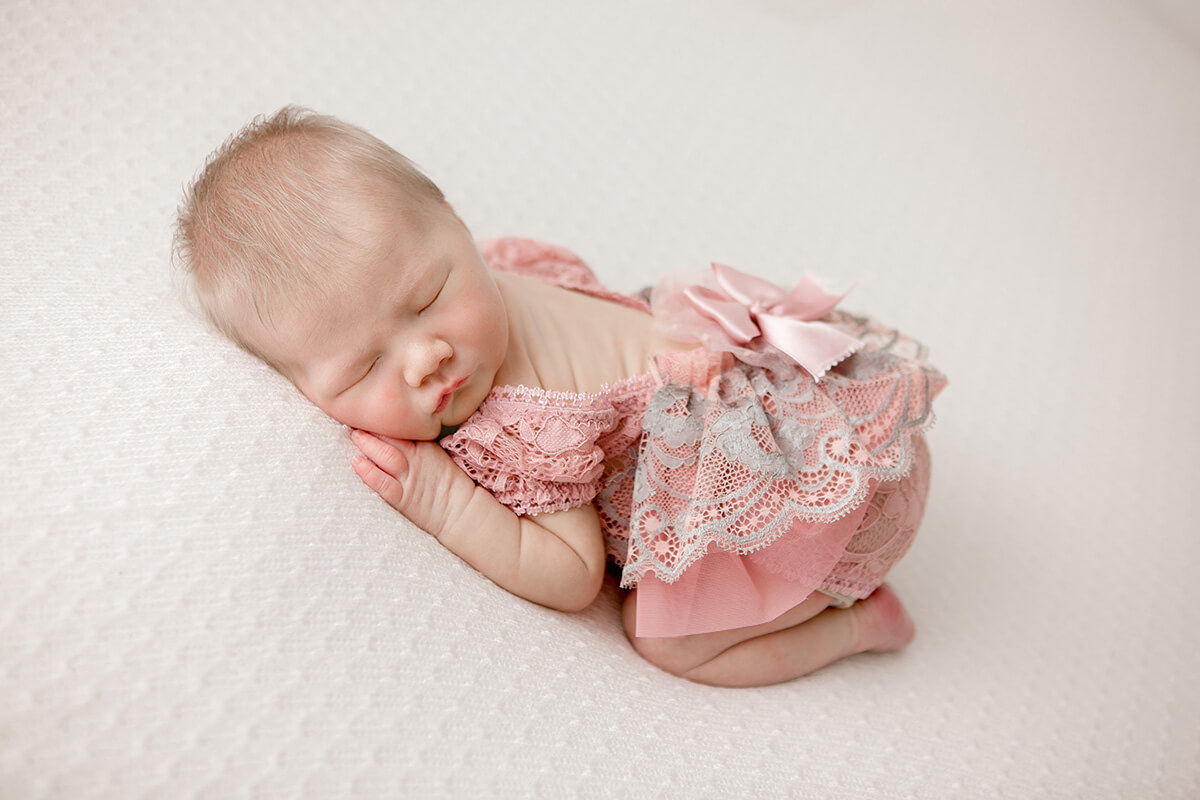 A baby girl in a pink lace dress sleeps in froggy pose on a white bed