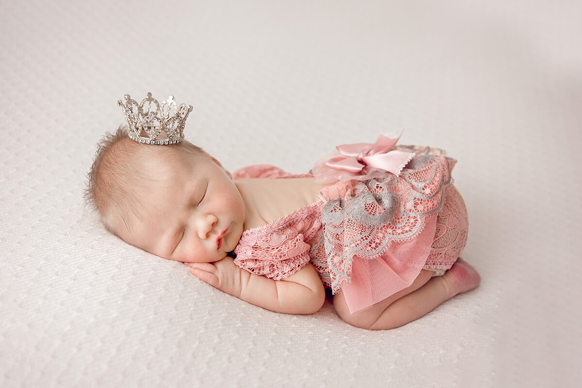 A newborn girl in a pink lace dress and tiny crown sleeping in froggy pose