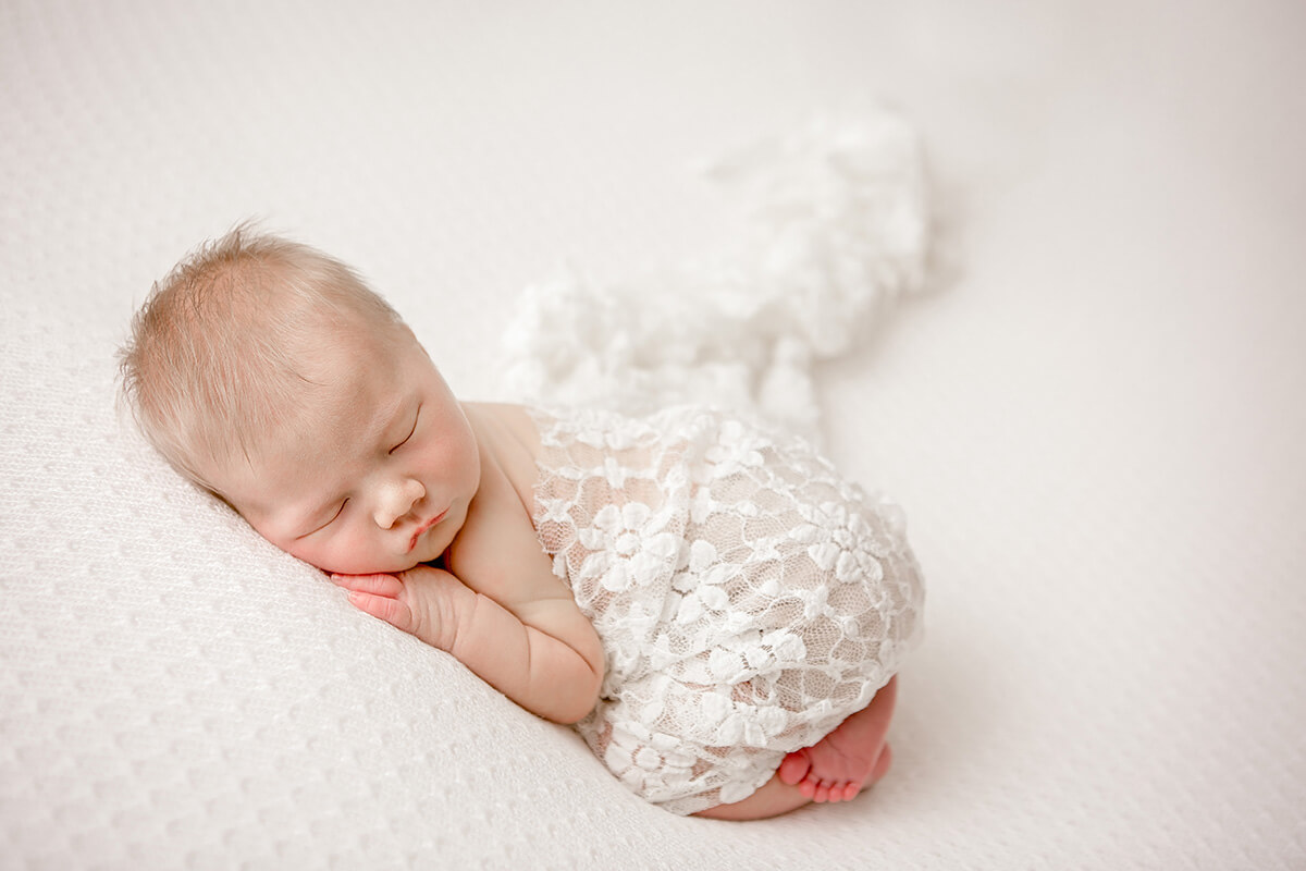 A newborn baby sleeps wrapped in a lace blanket on a white bed thanks to a midwife in bloomington il