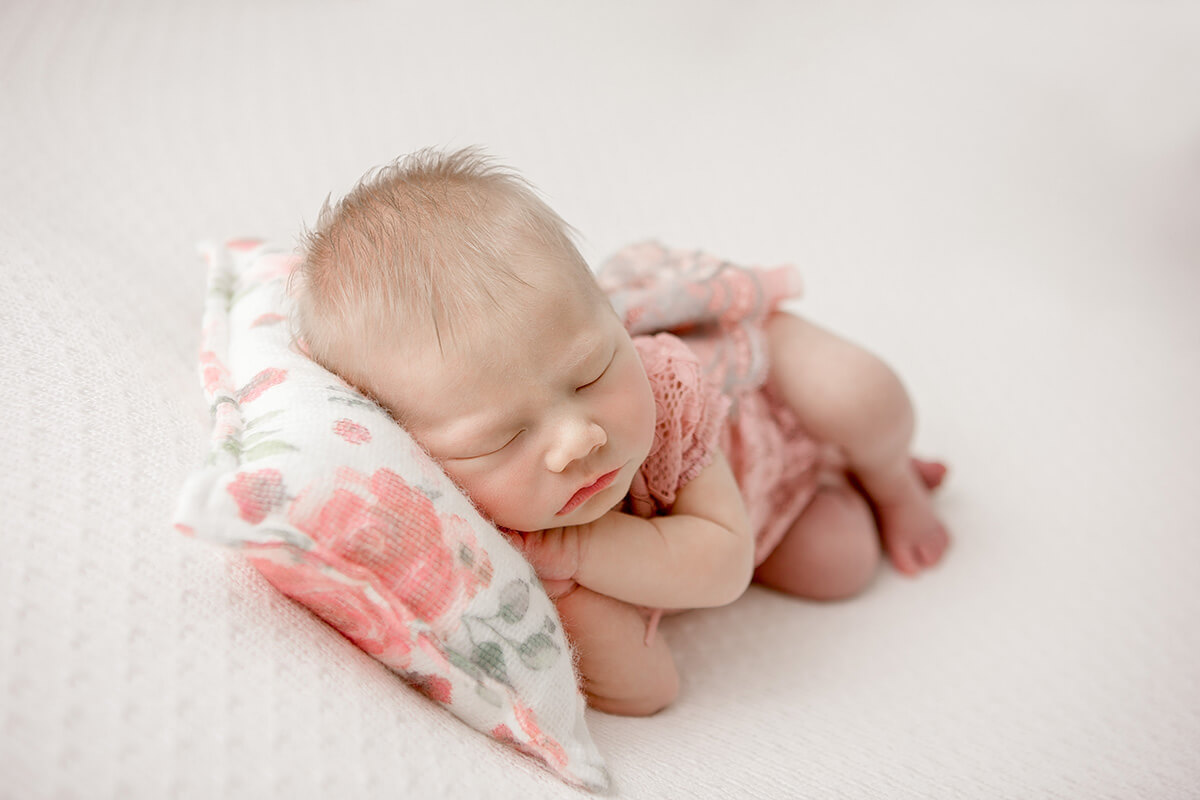 a newborn baby girl sleeps on a flower print pillow on a whtie bed in a pink lace dress thanks to a midwife in bloomington il