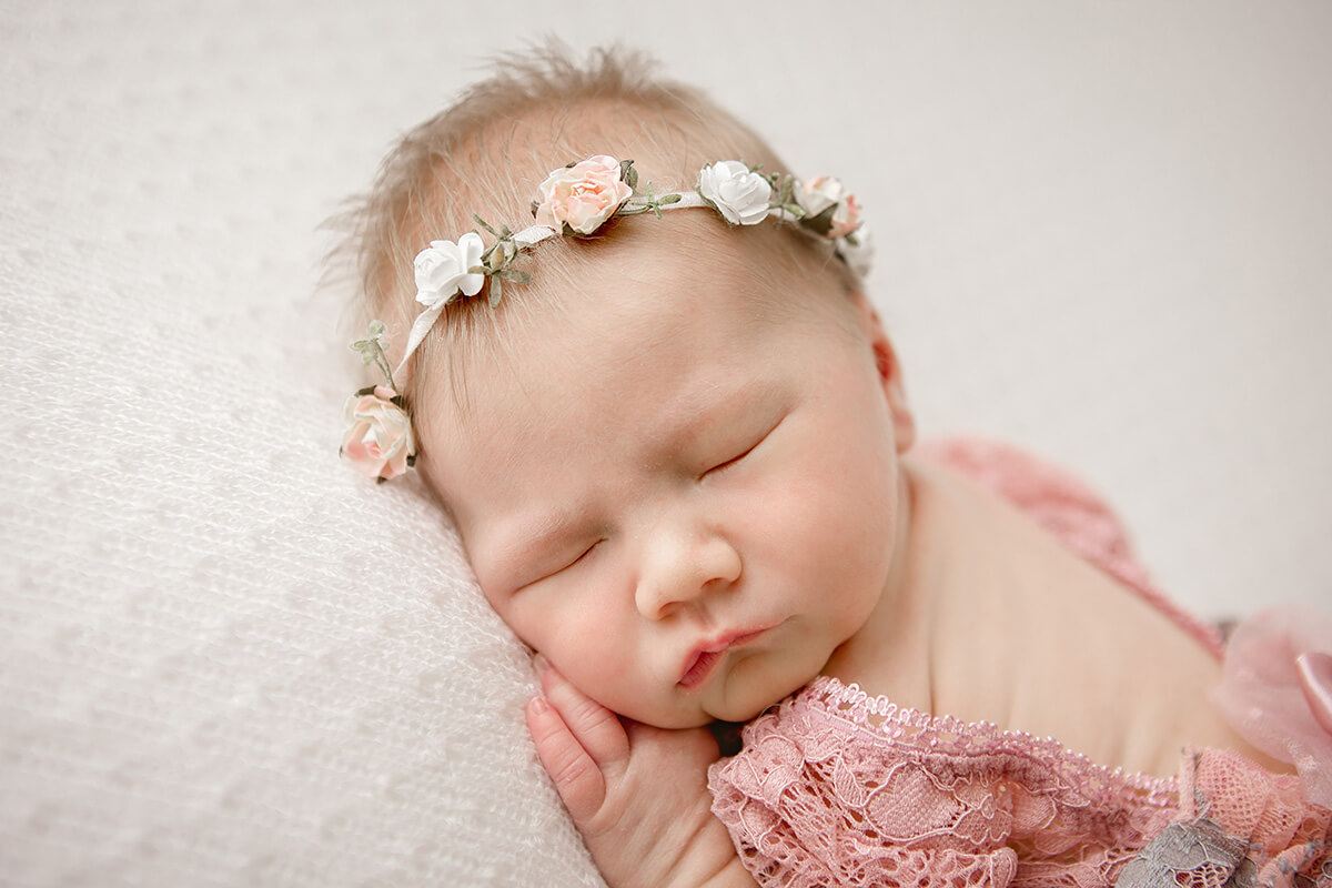 A sleepingn newborn on her tummy in a floral headband and pink lace gown