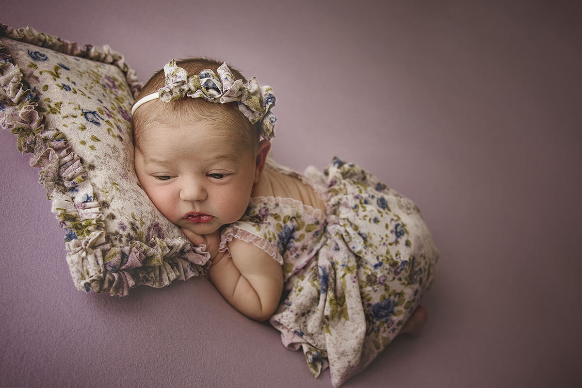 A baby girl in a floral print dress with eyes open on a purple bed resting on a matching pillow after mom found pelvic floor therapy in springfield, il