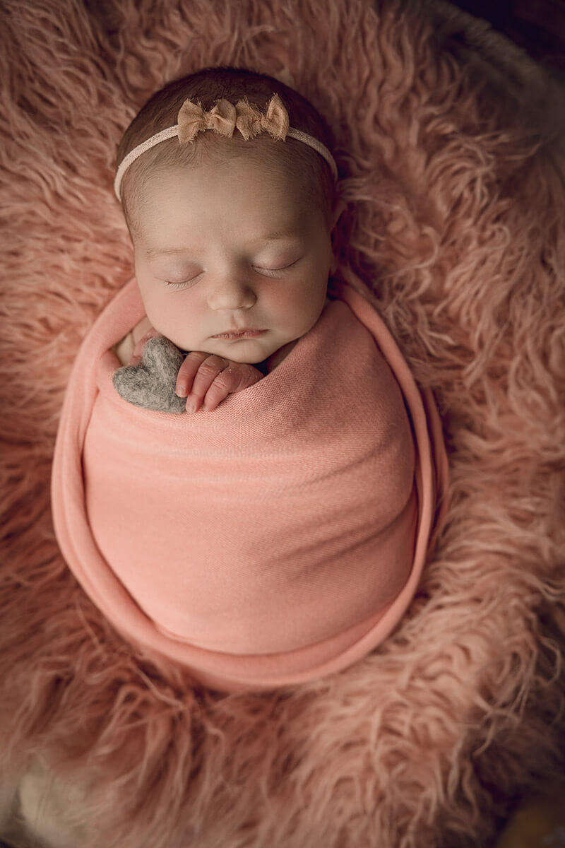 A sleeping newborn girl in a pink swaddle on a matching fur blanket holding a felt heart after mom found pelvic floor therapy in springfield, il