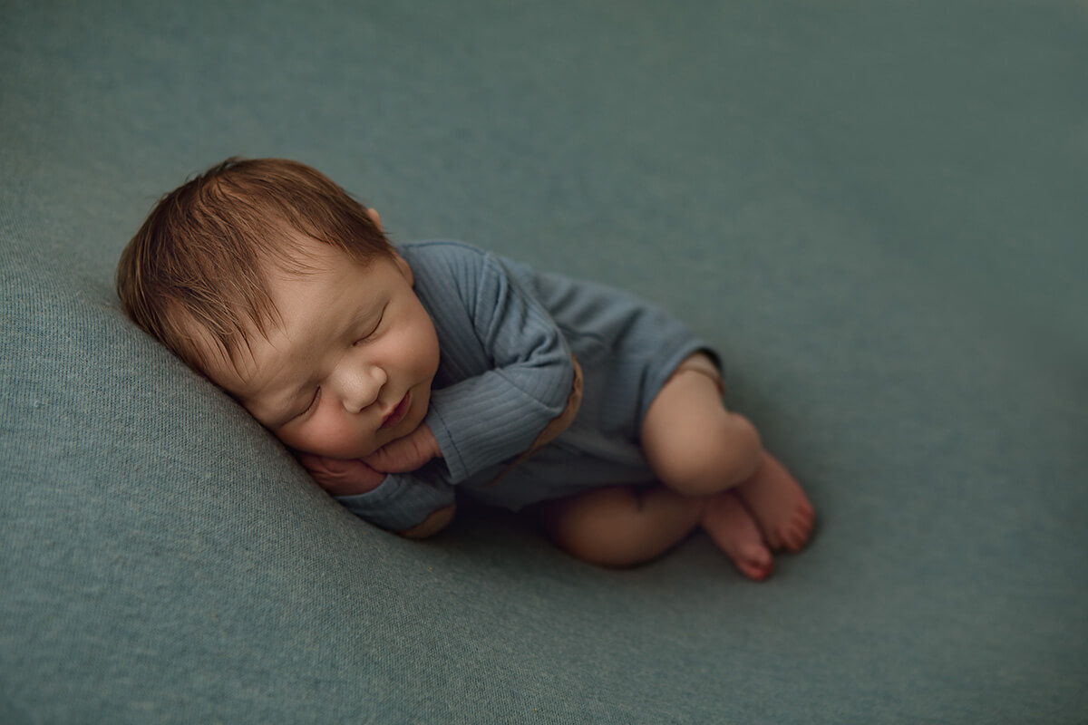A newborn baby sleeping on his side in a blue onesie with hands under his cheek