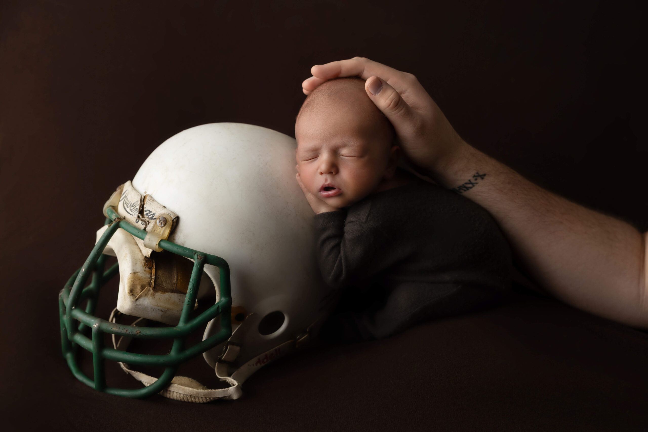 A dad holds the head of his newborn sleeping on an old football helmet after visiting baby shower venues in bloomington il