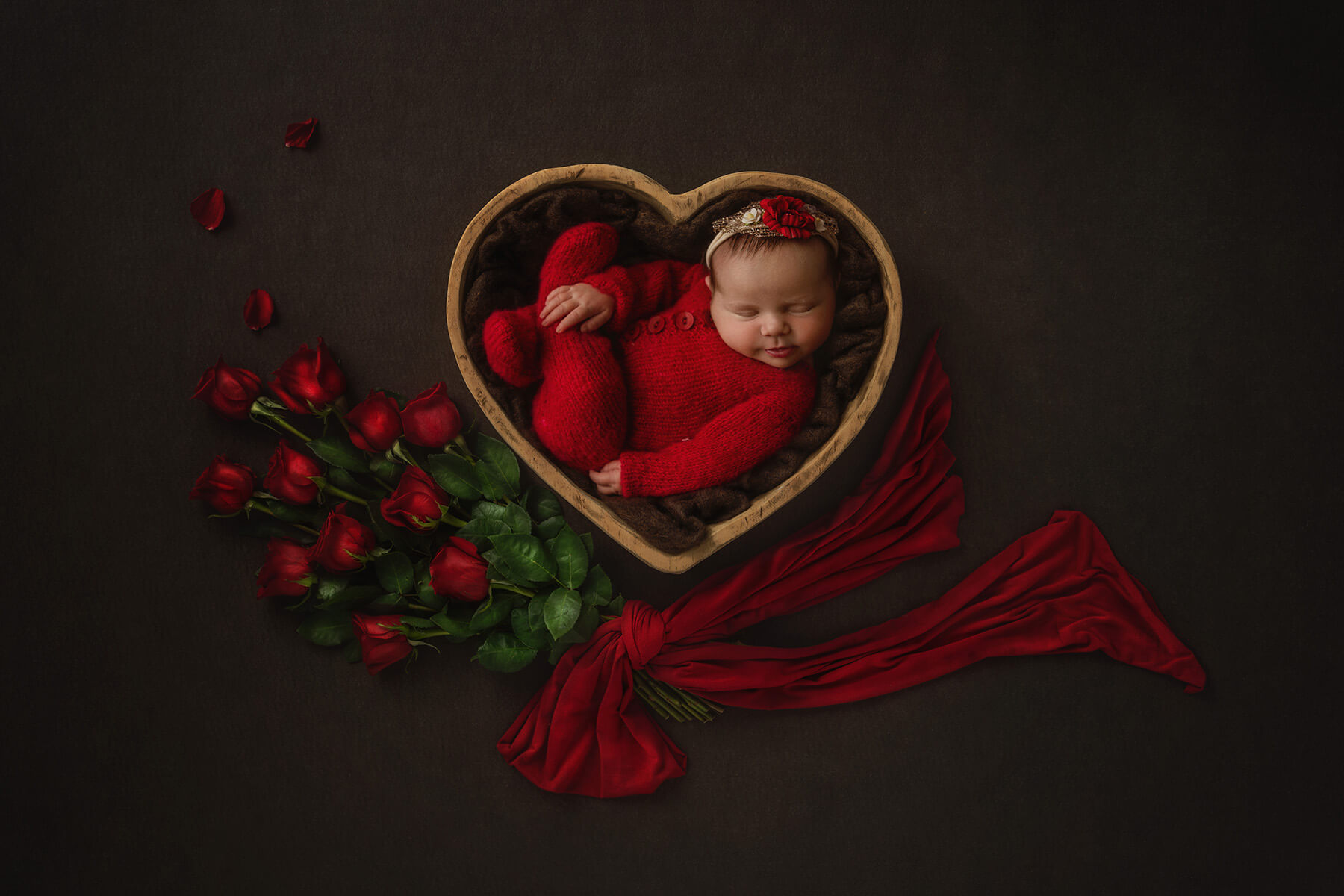 A newborn baby girl in a red knit onesie sleeps in a heart shaped bowl with a bouquet of red roses