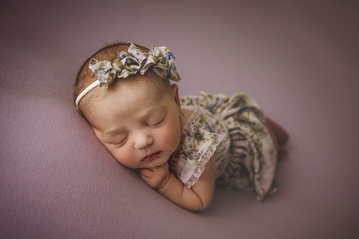 A newborn baby girl in a floral print dress and matching headband sleeps on a purple pad on her tummy thanks to ivf in Springfield, IL