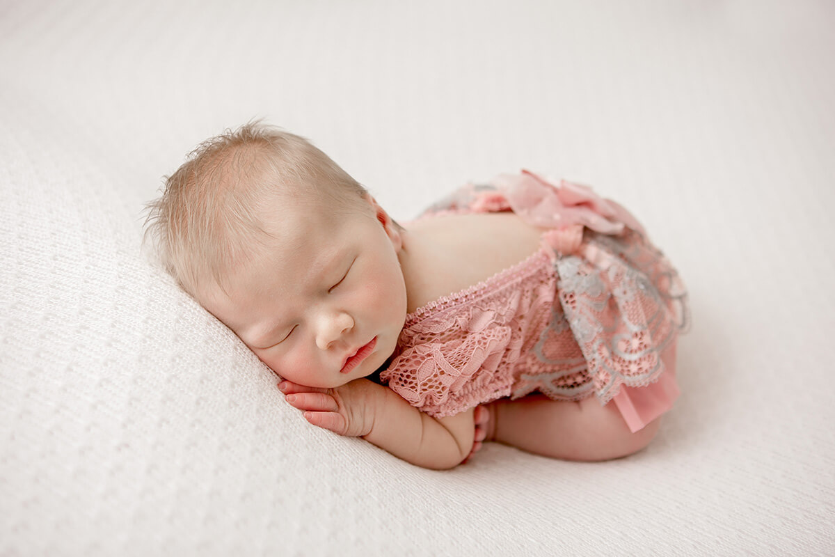 A newborn baby sleeps on her tummy in a pink lace onesie dress on a white bed